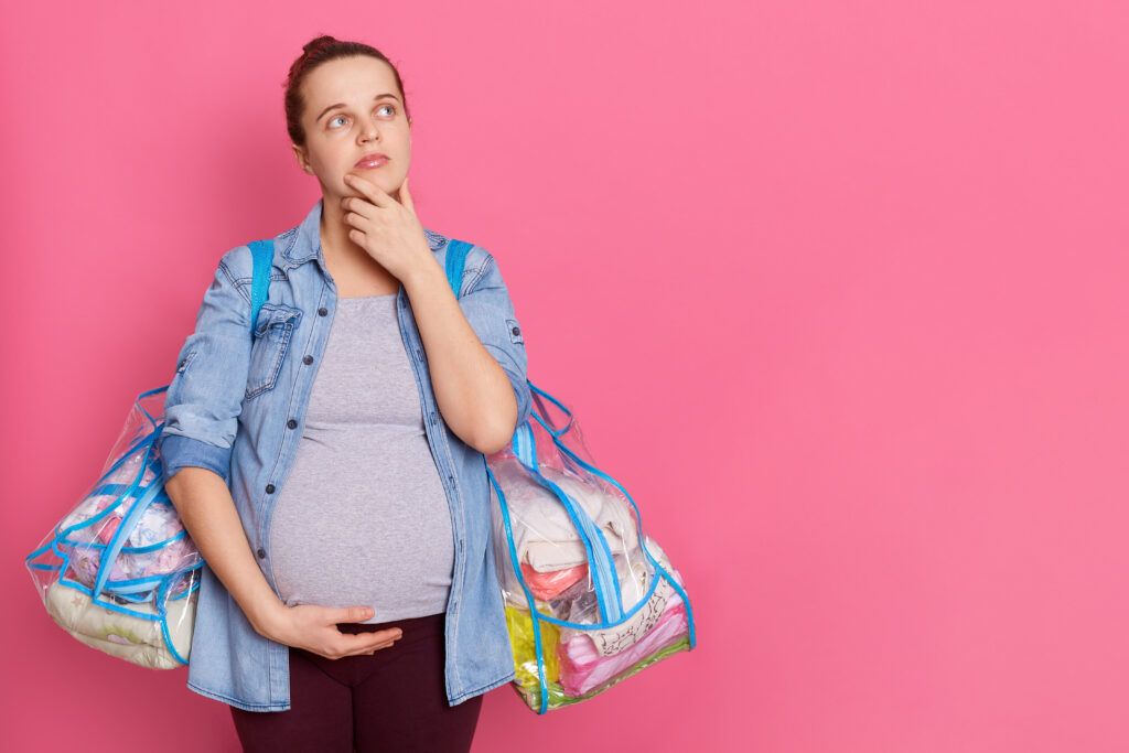 pregnant woman thinking while standing isolated over pink background, looking away and keeps palm on chin, wearing casual attire, touching her belly, holding two bags with stuff, copy space for ad.